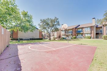 A basketball court is located in the middle of a grassy area in front of apartment buildings.